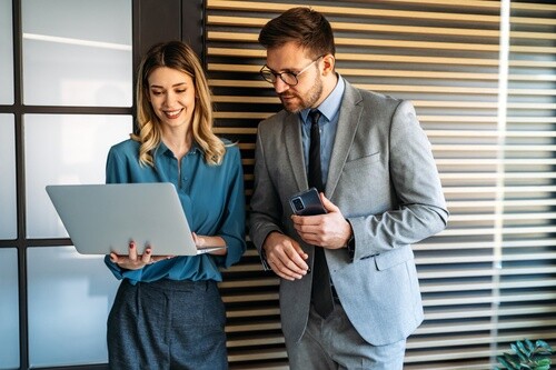 Two business professionals reviewing laptop screen while standing in office with smartphone